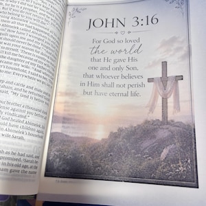 May include: Open Bible with a framed image of a cross on a hill, with the text "JOHN 3:16" and a quote about God's love. The image is set against a soft, sunset-like background.