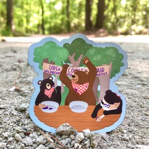 May include: Three cartoon bears wearing red and white checkered bandanas are sitting at a table with bowls of pie in front of them. The bears are participating in a pie eating contest at the Ursa Wildbear County Fair.