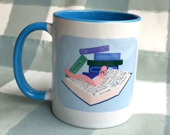 Puede incluir: Una taza de cerámica blanca con un borde y un asa azules. La taza presenta una ilustración de dibujos animados de un gusano con gafas leyendo un libro encima de una pila de libros. El texto "1011" es visible en la parte superior de los libros.