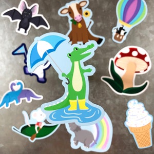 May include: A set of colorful stickers featuring a green alligator holding an umbrella, a cow, a mushroom, a hot air balloon, a dinosaur, a mouse, and a rainbow.