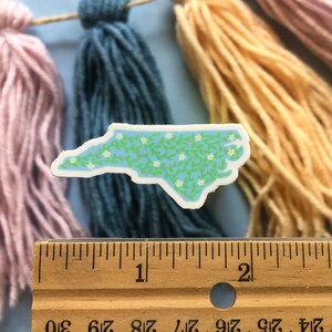 Puede incluir: Una pegatina de vinilo azul claro del estado de Carolina del Norte con un patr&oacute;n de enredadera verde y flores blancas. La pegatina mide aproximadamente 5 cm de ancho.