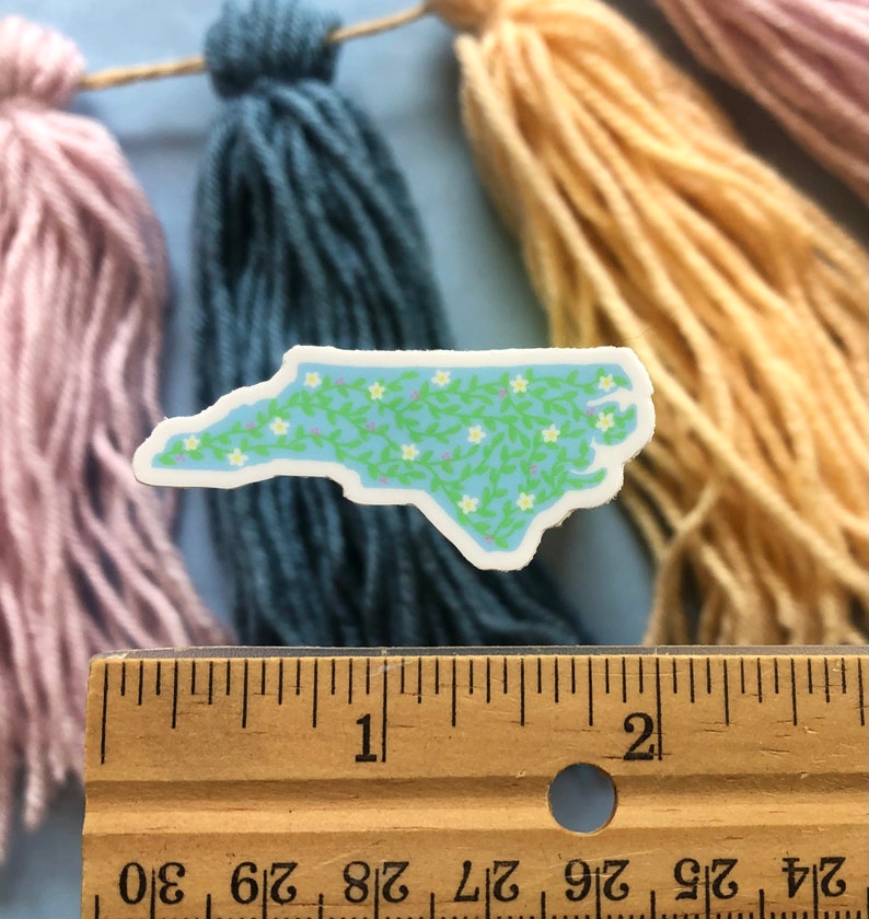 Puede incluir: Una pegatina de vinilo azul claro del estado de Carolina del Norte con un patr&oacute;n de enredadera verde y flores blancas. La pegatina mide aproximadamente 5 cm de ancho.