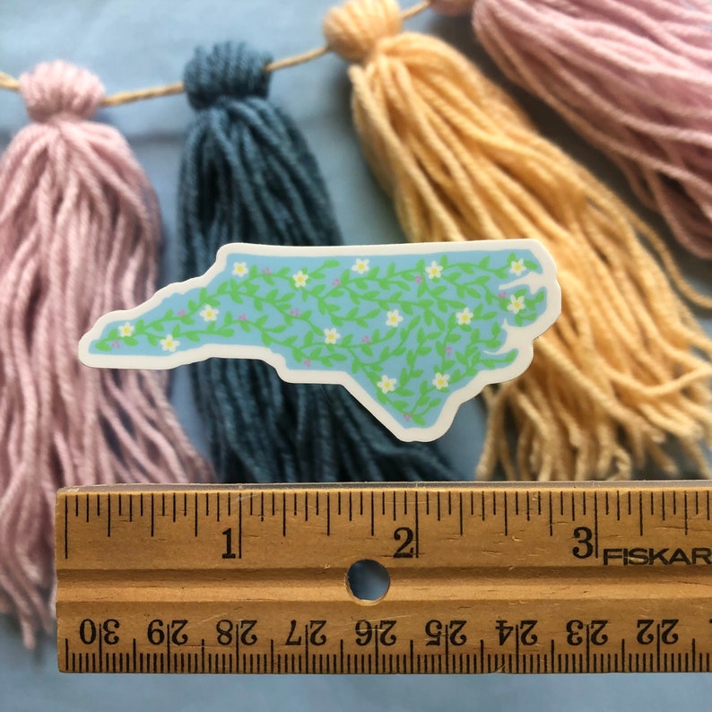 Puede incluir: Una pegatina de vinilo azul y verde del estado de Carolina del Norte con flores blancas y enredaderas verdes. La pegatina mide aproximadamente 5 cm de largo y est&aacute; sobre una regla de madera.