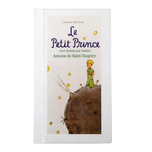 May include: A hardcover book titled "Le Petit Prince" by Antoine de Saint-Exupéry. The cover features an illustration of a young boy in green clothing standing on a small planet, with stars and a yellow moon in the background.