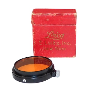 May include: A vintage Leica camera filter with a black metal frame and an orange-tinted glass lens. The filter sits next to a red box with the Leica logo and the text "E. Leitz, Inc. New York."