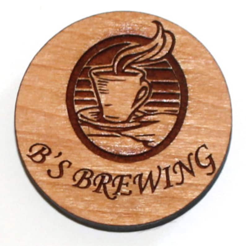 Puede incluir: Un posavasos redondo de madera con un dise&ntilde;o grabado en marr&oacute;n de una taza de caf&eacute; humeante. El texto "B'S BREWING" est&aacute; grabado debajo del dise&ntilde;o.
