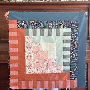 May include: A square quilt featuring a log cabin block design with a central light pink and floral fabric square. It is surrounded by borders of sage green, dark blue with a forest animal print, and a rust-coloured striped fabric border.
