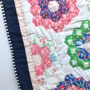 May include: Close-up of a handmade quilt with a floral pattern. The quilt features a patchwork design with hexagonal shapes in pink, blue, and green fabrics. A navy blue border with white stripes and a light pink trim complete the design.