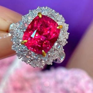 May include: A ring with a large, vibrant red gemstone surrounded by a halo of smaller, sparkling white gemstones.