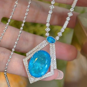 May include: A silver necklace featuring a large, oval, light blue gemstone pendant. The pendant is surrounded by a geometric frame and small, clear gemstones. The necklace chain is delicate and sparkly.