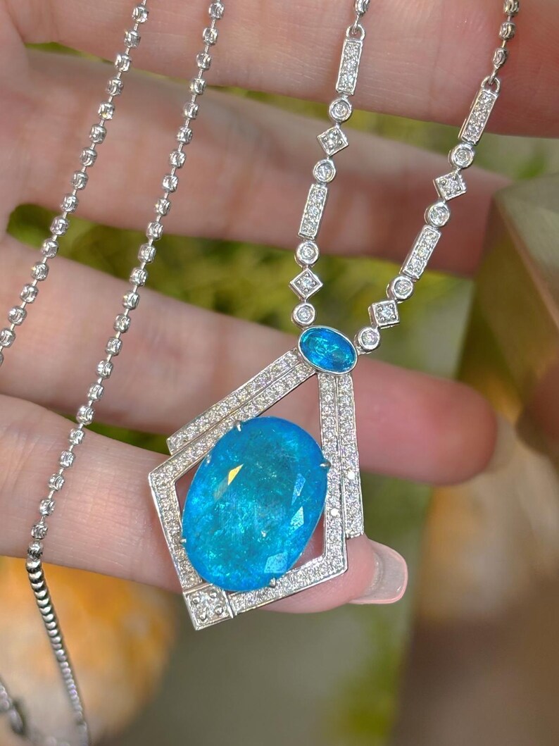 May include: A silver necklace featuring a large, oval, light blue gemstone pendant. The pendant is surrounded by a geometric frame and small, clear gemstones. The necklace chain is delicate and sparkly.