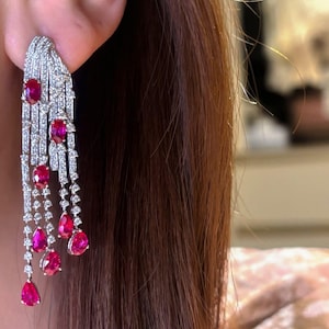 May include: Elegant chandelier earrings with a silver setting, featuring numerous small, sparkling clear stones and vibrant red ruby gemstones. The earrings have a cascading design with teardrop-shaped gems.