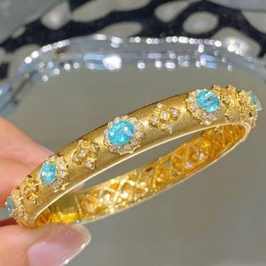 May include: A gold bangle bracelet with a brushed finish and a row of blue gemstones set in a diamond-shaped pattern.