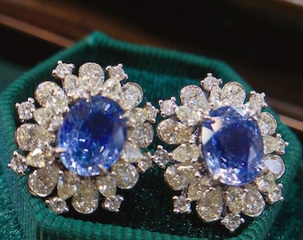 May include: A pair of earrings with a large blue sapphire gemstone surrounded by a halo of smaller diamonds. The earrings are set in a white metal.
