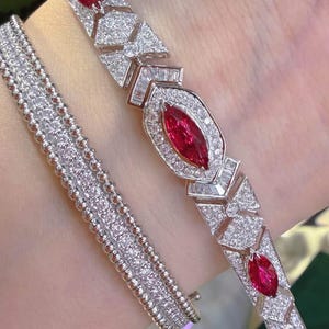 Könnte beinhalten: Ein silberfarbenes Armband mit marquise-geschliffenen roten Edelsteinen in geometrischen Fassungen, besetzt mit kleinen weißen Steinen, kombiniert mit einem zweiten Armband aus mehreren Reihen winziger silberner Perlen.
