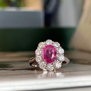May include: A silver ring featuring a vibrant pink oval gemstone, surrounded by a halo of smaller, sparkling white gemstones. The ring is set against a blurred background, highlighting the jewelry's details.