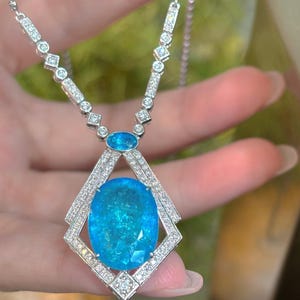 May include: A silver necklace featuring a large, oval, light blue gemstone pendant. The pendant is surrounded by a diamond-studded frame. The necklace chain is also adorned with diamonds and smaller blue gemstones, creating a luxurious and elegant design.