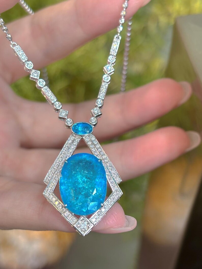 May include: A silver necklace featuring a large, oval, light blue gemstone pendant. The pendant is surrounded by a diamond-studded frame. The necklace chain is also adorned with diamonds and smaller blue gemstones, creating a luxurious and elegant design.