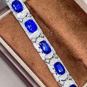 May include: A luxurious bracelet featuring large, oval-cut, deep blue gemstones set in a silver or white gold band. The bracelet is adorned with smaller, clear gemstones, creating a sparkling contrast. The jewelry is displayed in a brown jewelry box.