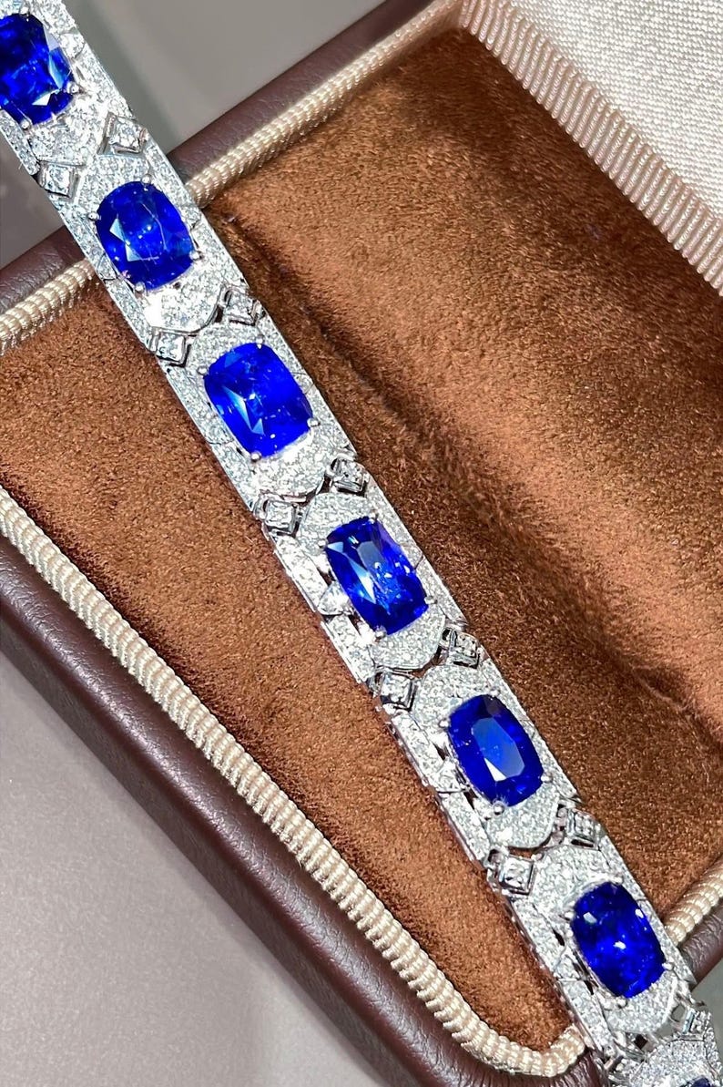 May include: A luxurious bracelet featuring large, oval-cut, deep blue gemstones set in a silver or white gold band. The bracelet is adorned with smaller, clear gemstones, creating a sparkling contrast. The jewelry is displayed in a brown jewelry box.