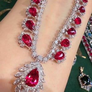 May include: A silver-toned necklace featuring a series of pear-shaped red gemstones set in metal frames surrounded by small clear stones. A large, central pear-shaped red gemstone pendant is framed by marquise-cut clear stones in a leaf-like pattern.