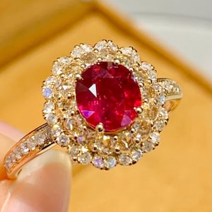 May include: A gold ring featuring a large, oval-cut ruby gemstone surrounded by a halo of smaller diamonds. The ring's band is also adorned with small diamonds, creating a sparkling and luxurious appearance.