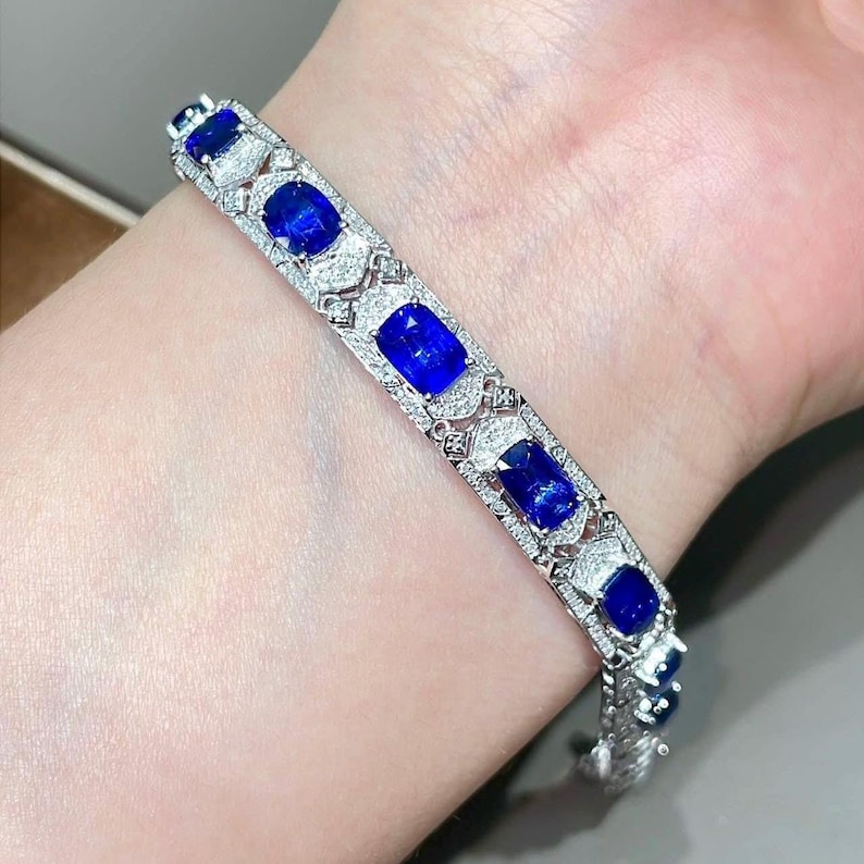 May include: A silver bracelet with multiple oval and rectangular blue gemstones, interspersed with smaller clear stones. The bracelet is set with intricate filigree detailing, creating a luxurious and elegant appearance. The gemstones are a vibrant blue color.