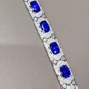 May include: A close-up of a silver bracelet featuring large, oval-cut, deep blue gemstones. Each gemstone is surrounded by a halo of smaller, sparkling white stones. The bracelet is set against a neutral background.