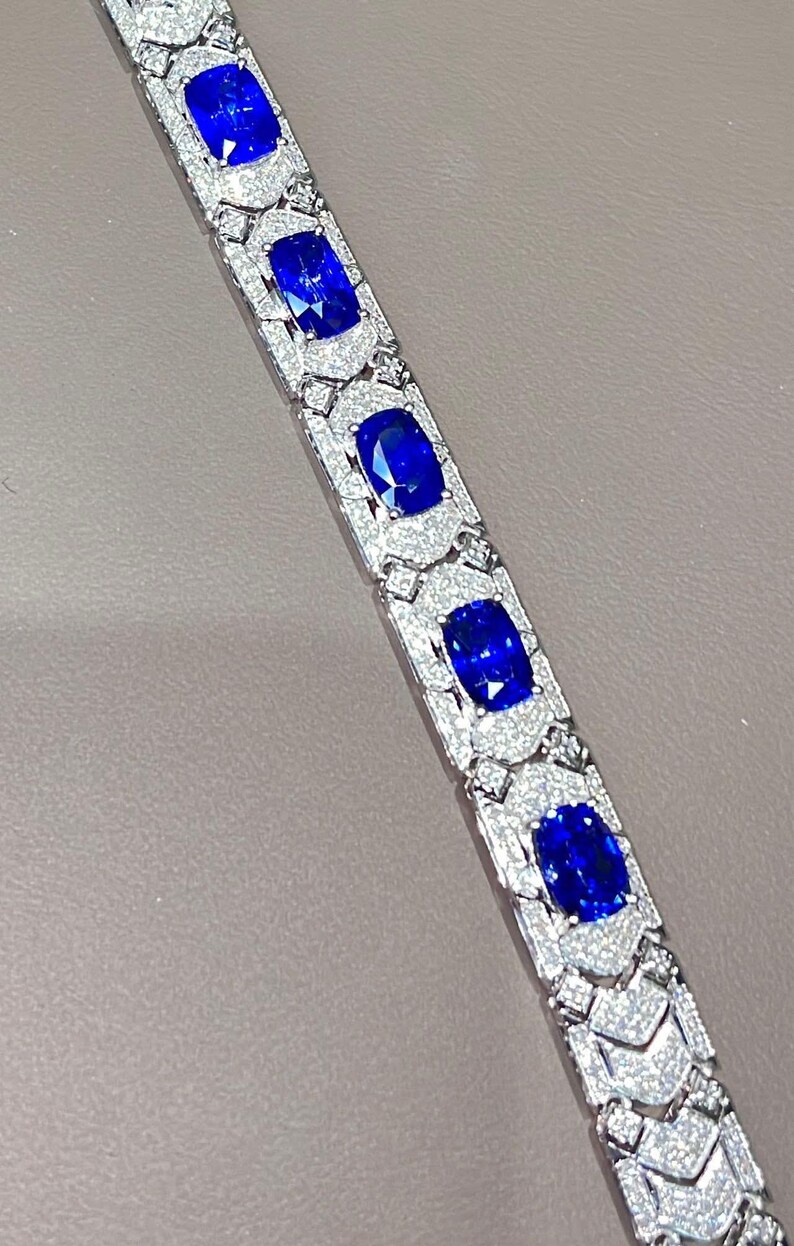 May include: A close-up of a silver bracelet featuring large, oval-cut, deep blue gemstones. Each gemstone is surrounded by a halo of smaller, sparkling white stones. The bracelet is set against a neutral background.