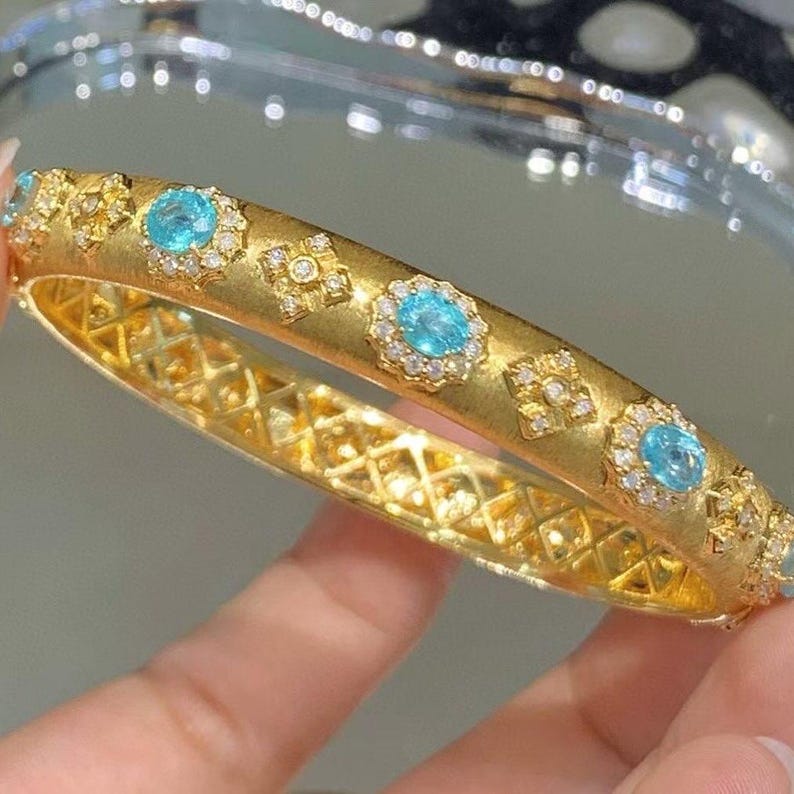 May include: A gold bangle bracelet with a delicate, intricate design. The bracelet is adorned with small, round blue gemstones and sparkling diamonds.