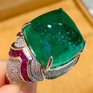 May include: A striking ring featuring a large, emerald-cut green gemstone. The ring has a square-cut emerald, complemented by smaller rubies and diamonds set in a silver band. A bold statement piece.
