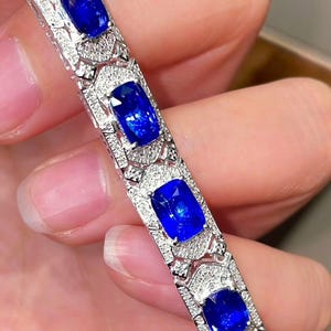 May include: A close-up of a silver bracelet featuring large, rectangular, deep blue gemstones. The bracelet is adorned with smaller, clear gemstones around the larger stones, creating a sparkling effect. The design is intricate and elegant.