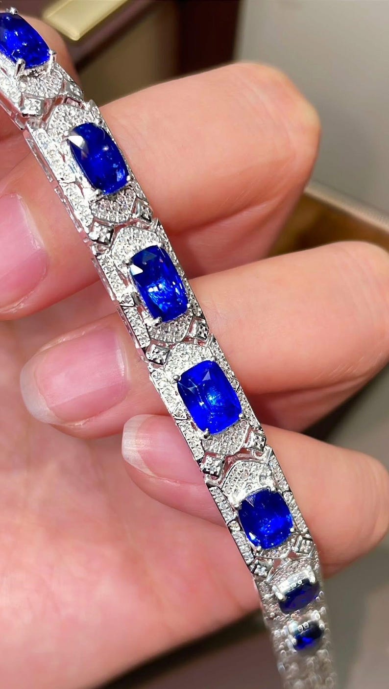 May include: A close-up of a silver bracelet featuring large, rectangular, deep blue gemstones. The bracelet is adorned with smaller, clear gemstones around the larger stones, creating a sparkling effect. The design is intricate and elegant.