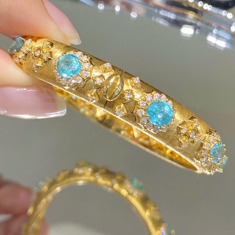 May include: A gold bangle bracelet with a textured surface and a row of blue gemstones set in a halo of smaller white gemstones.