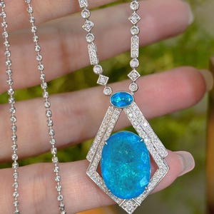 May include: A silver necklace featuring a large, oval, teal-colored gemstone pendant. The pendant is framed by a diamond-studded geometric design. The necklace chain is delicate, with small, round links. Additional small gemstones are set along the chain.