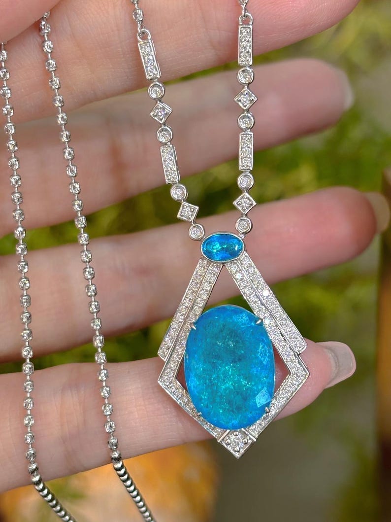 May include: A silver necklace featuring a large, oval, teal-colored gemstone pendant. The pendant is framed by a diamond-studded geometric design. The necklace chain is delicate, with small, round links. Additional small gemstones are set along the chain.