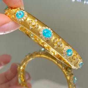 May include: A pair of gold bangles with a detailed design featuring turquoise gemstones and small diamonds. The bangles are stacked on top of each other.