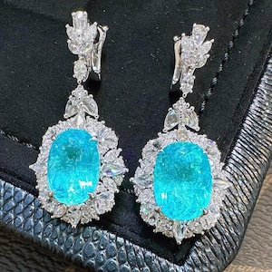 May include: A pair of silver drop earrings featuring a large oval turquoise gemstone surrounded by smaller clear gemstones.