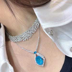 May include: A two-strand diamond necklace with a large, oval, bright blue gemstone pendant. The upper necklace is a wide band of diamonds. The pendant is set in a geometric frame, and the necklace is worn over a white shirt.