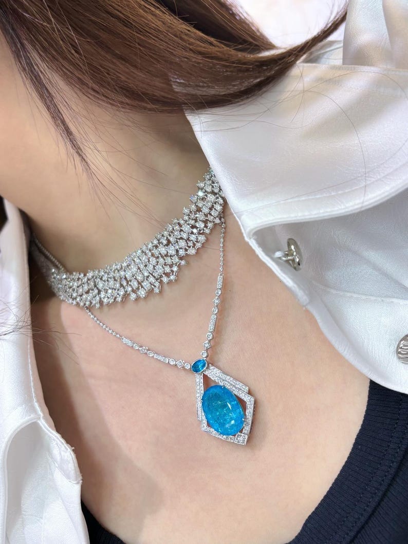 May include: A two-strand diamond necklace with a large, oval, bright blue gemstone pendant. The upper necklace is a wide band of diamonds. The pendant is set in a geometric frame, and the necklace is worn over a white shirt.