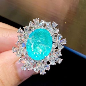 May include: A turquoise gemstone ring with a floral design. The ring is set in silver and features a large oval-shaped gemstone surrounded by smaller diamonds.