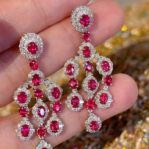 以下が含まれることがあります： 鮮やかな赤い宝石が連なった、揺れるタイプのイヤリング。それぞれの宝石は、輝く白いダイヤモンドの光輪で囲まれています。イヤリングはクラスターデザインで、さまざまなサイズの複数の宝石が使用されています。