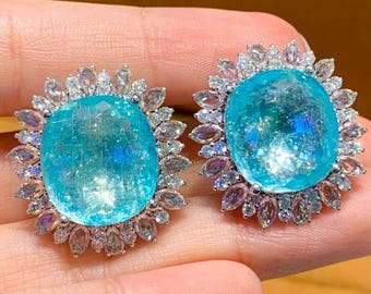May include: Two striking earrings, each with a large, oval, light blue gemstone at the centre. These are encircled by a halo of smaller, clear gemstones. The earrings are set in a silver-toned metal, creating a floral-like design.
