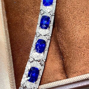 May include: A close-up of a silver bracelet with large, square, deep blue gemstones. The bracelet is encrusted with smaller, clear gemstones. The bracelet is displayed in a brown jewelry box.