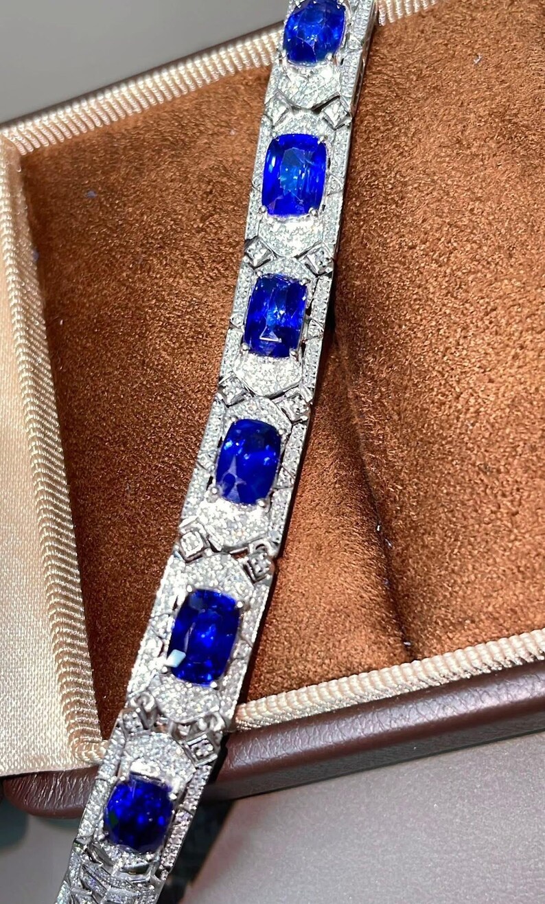 May include: A close-up of a silver bracelet with large, square, deep blue gemstones. The bracelet is encrusted with smaller, clear gemstones. The bracelet is displayed in a brown jewelry box.
