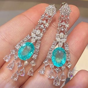 May include: A pair of drop earrings with large, oval, teal gemstones. The earrings are embellished with smaller, clear gemstones and detailed metalwork. The design is intricate and sparkling, creating a luxurious appearance.
