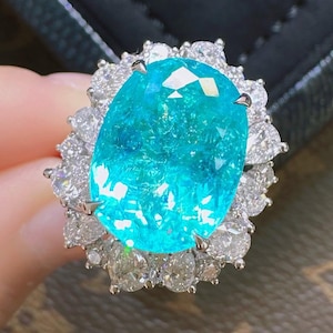 May include: A striking ring with a large, oval-cut, bright turquoise gemstone at its centre. The gemstone is encircled by a halo of smaller, clear, pear-shaped diamonds, creating a dazzling display of light and colour. The ring's setting appears to be silver or white gold.