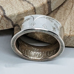 May include: A silver half dollar coin ring with the year 1965 and the words "In God We Trust" engraved on the band.