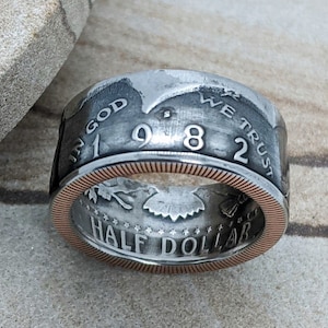 May include: A silver half dollar coin ring with a copper band. The coin is dated 1982 and has the inscription "In God We Trust" and "Half Dollar".