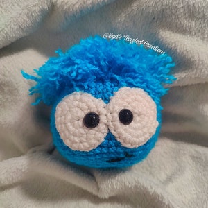 May include: A blue crocheted plush toy with white eyes and a fuzzy blue hair. The toy is on a white fluffy surface. @Syd's Tangled Creations